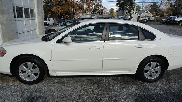 White four-door sedan parked on a gravel lot near a building and trees.