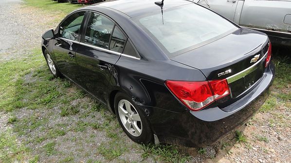 Black sedan parked on a gravel driveway beside grass, viewed from the rear-left side.