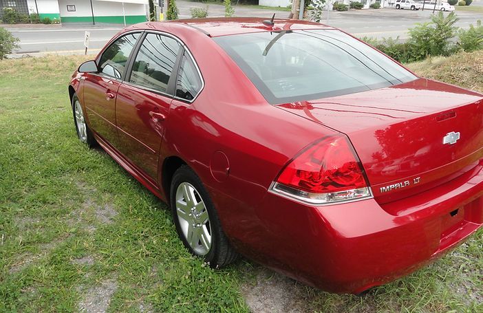 Red sedan parked on grass beside a road, rear three-quarter view.