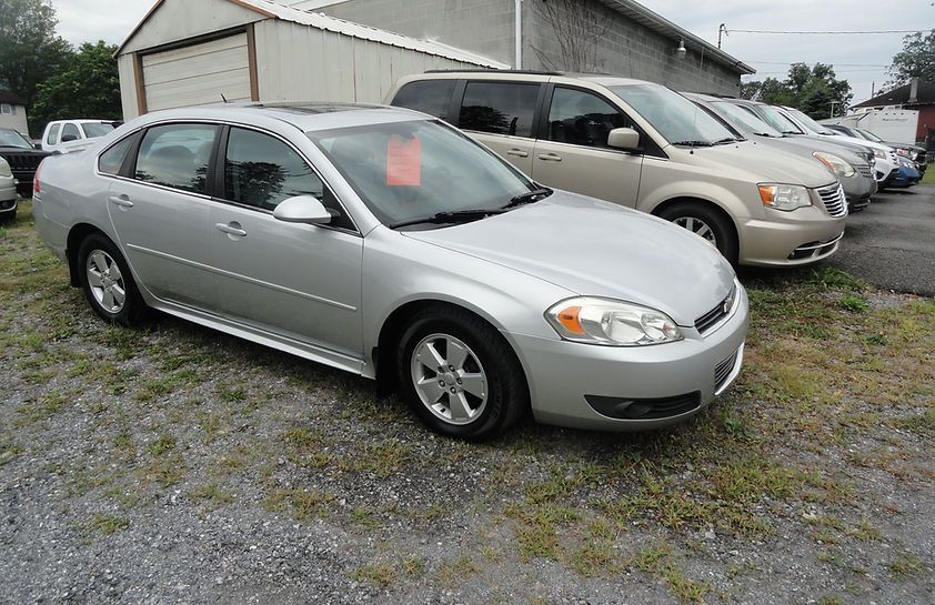 Silver sedan parked in a gravel lot beside beige SUVs and a metal building