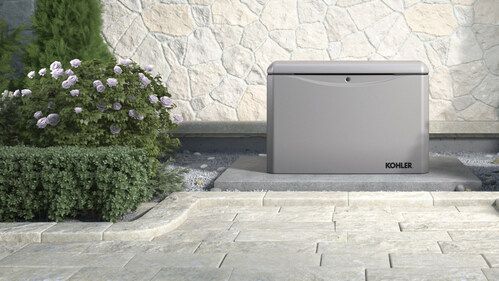 A gray outdoor generator on a concrete pad, next to a garden with a stone wall background.
