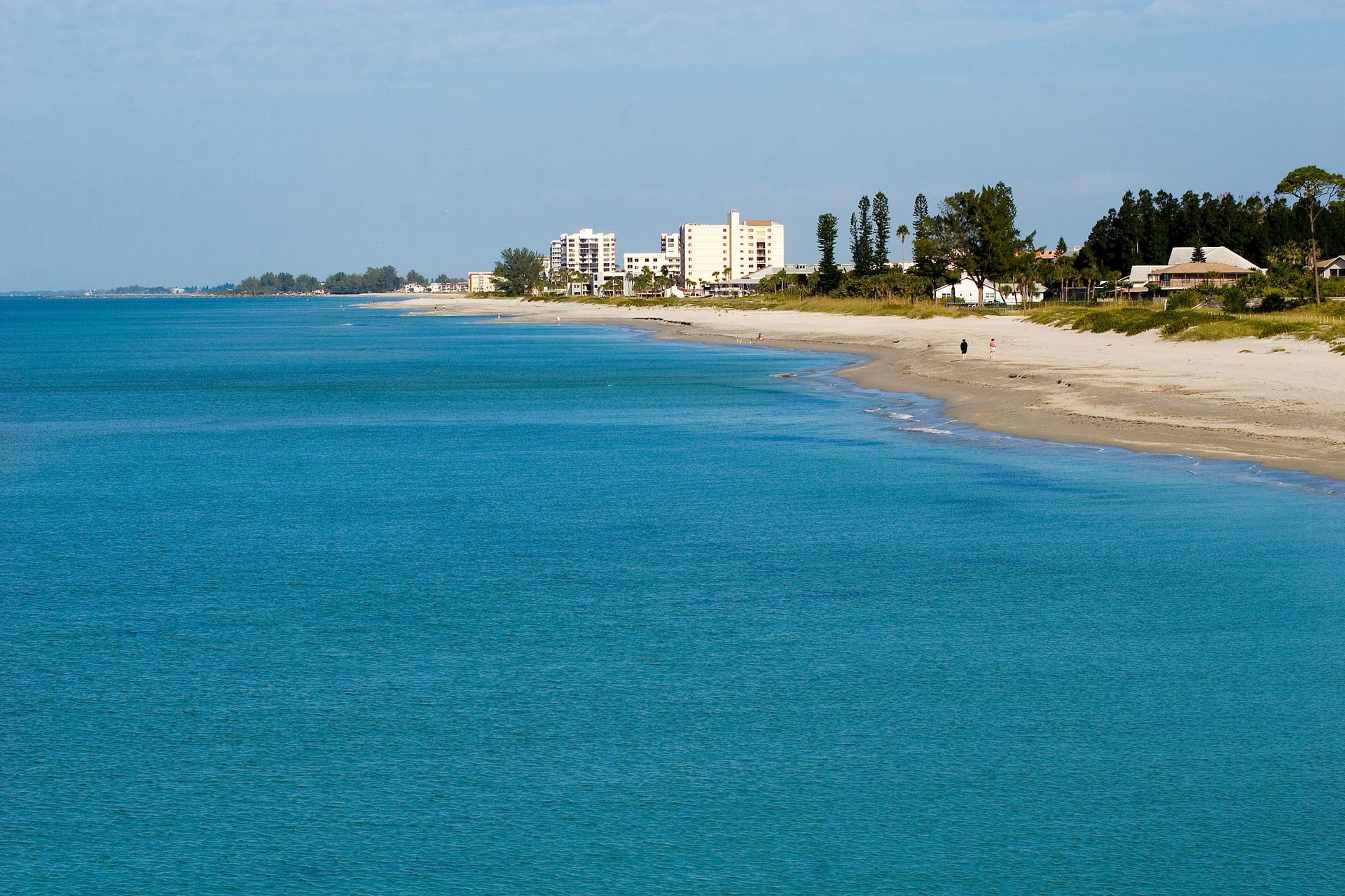 Bright blue sea meets sandy beach with white buildings and trees in the background under a blue sky.