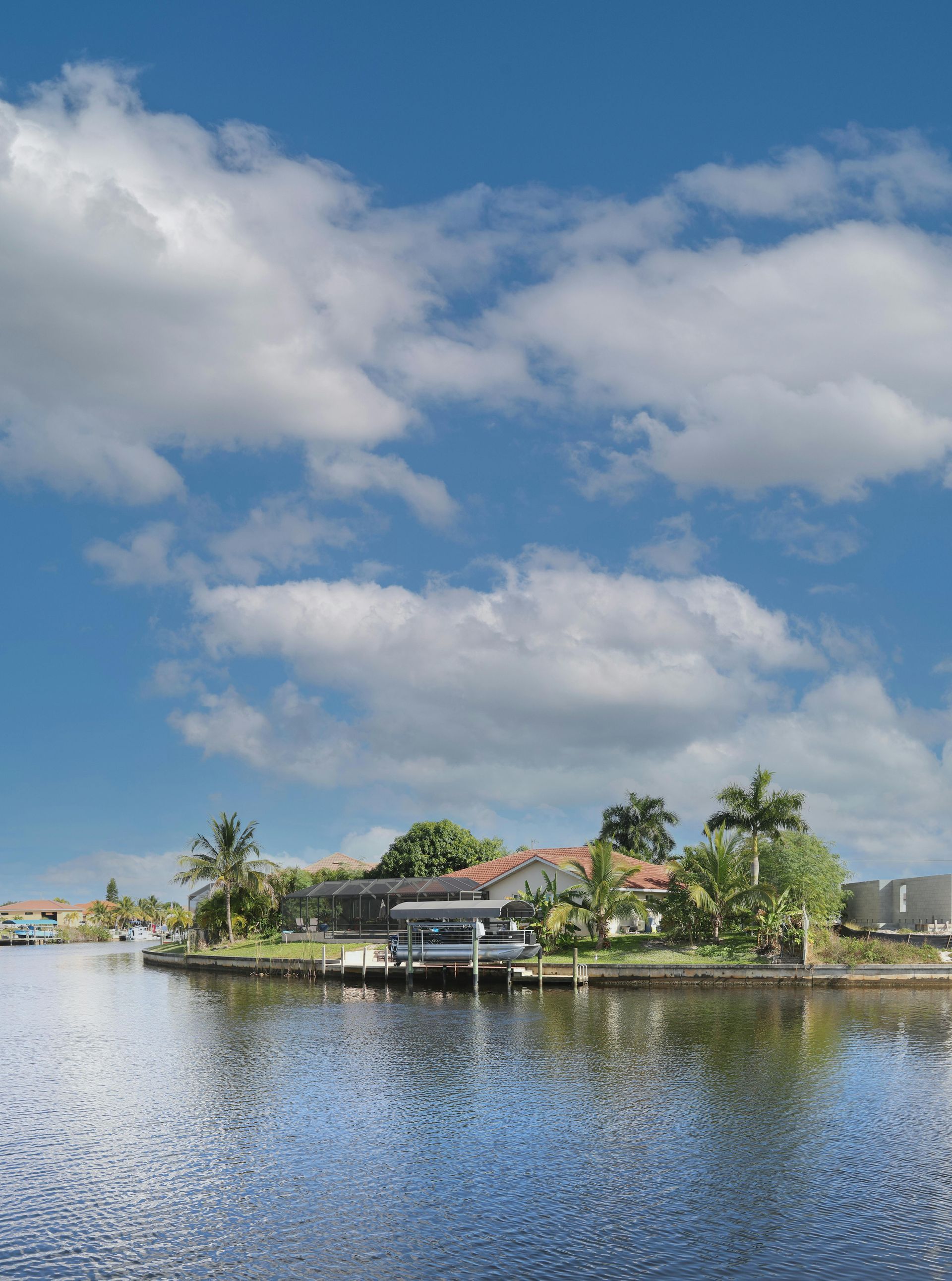 Waterfront house with dock, palm trees, and blue sky with puffy white clouds.