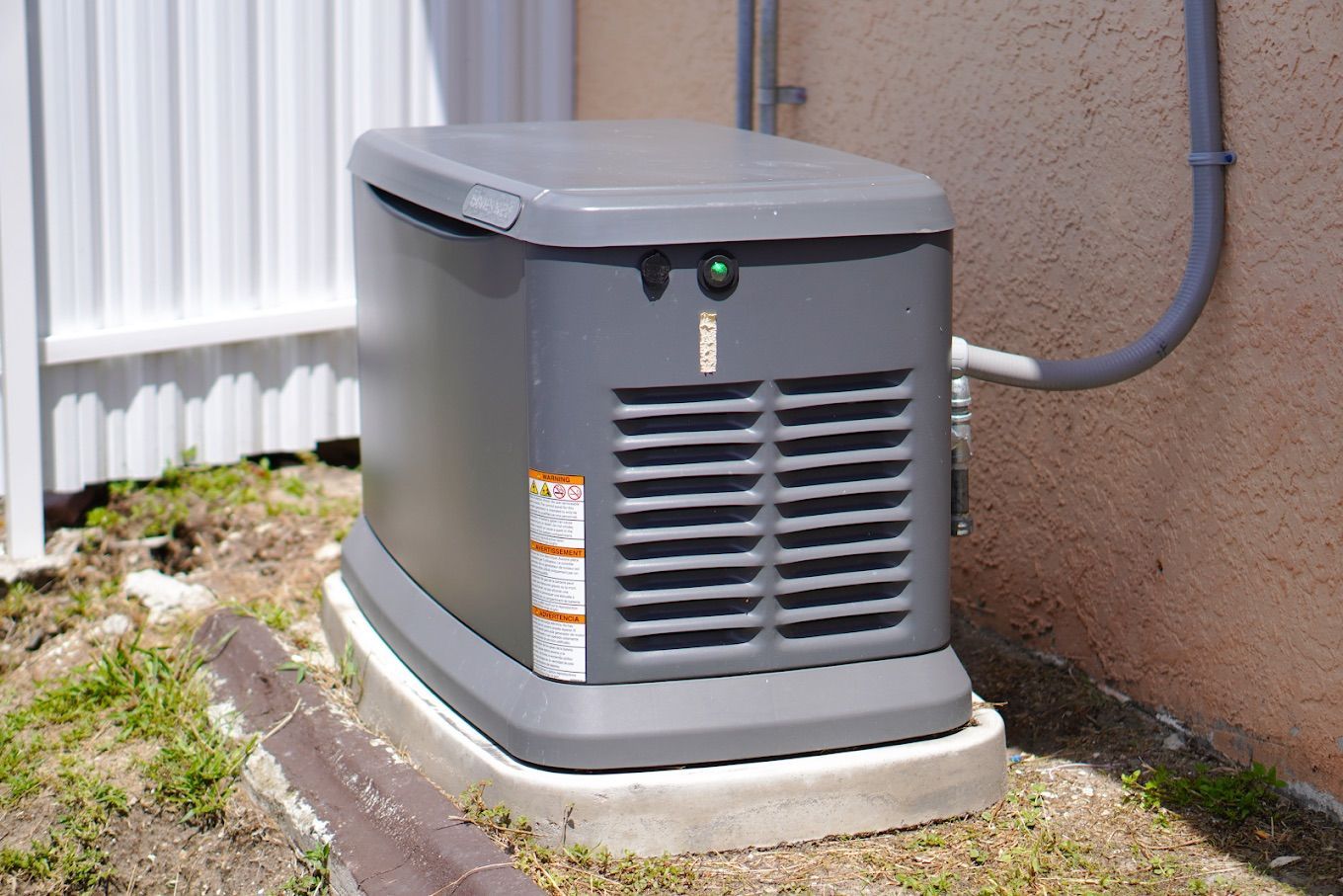 Gray home generator on a concrete base, near a white wall and tan building.