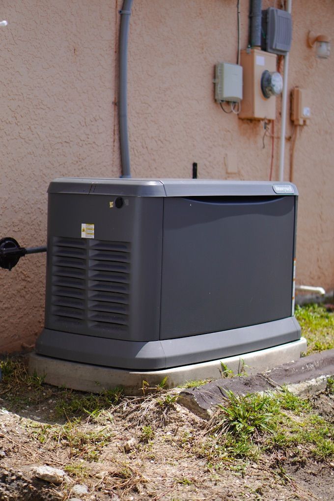 Dark gray generator outside, next to a beige building with electrical components.