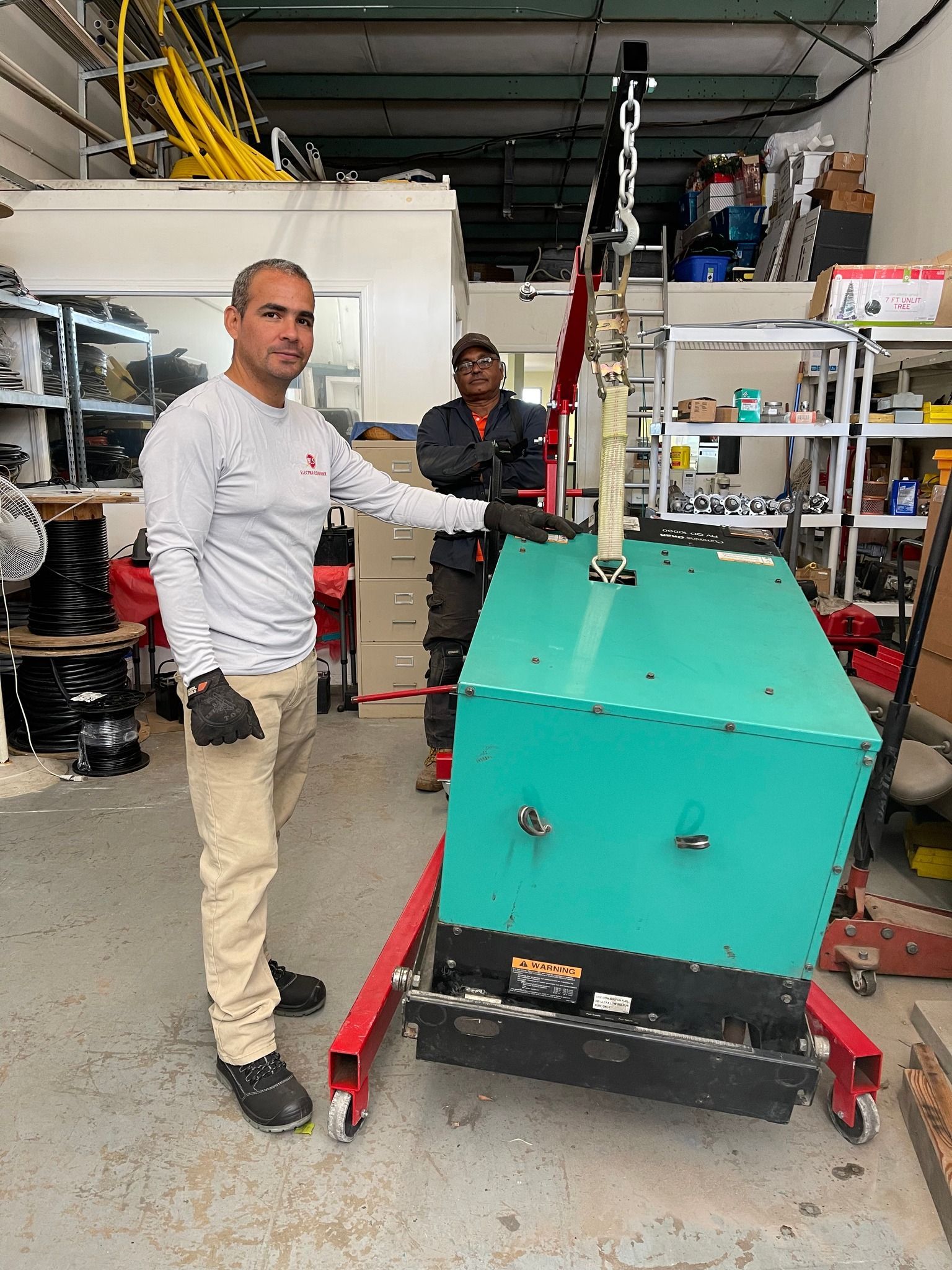 Two men in a workshop lift a large teal-colored generator with a hoist. One points at it.