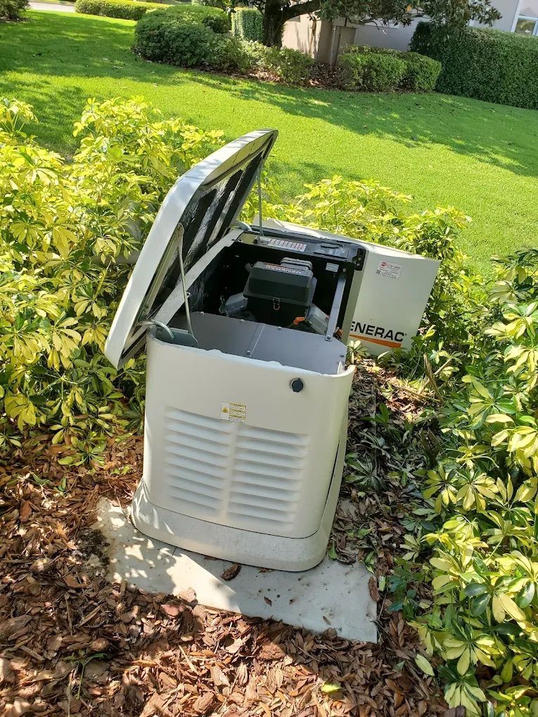 White and tan home generator with open lid in a yard, surrounded by green bushes.