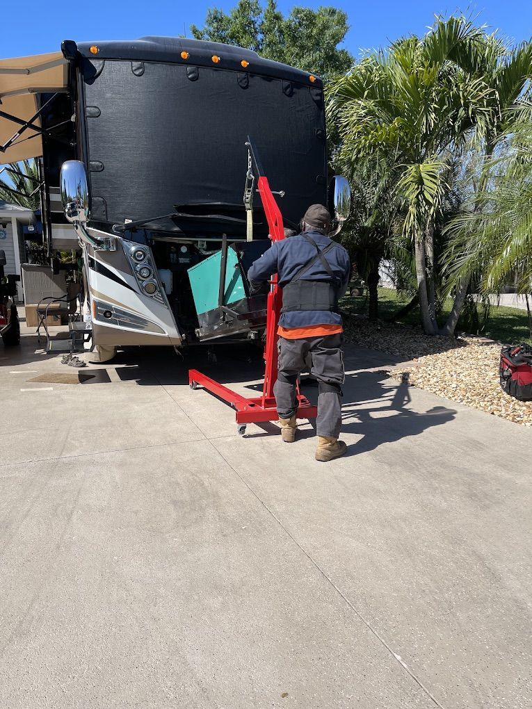 Person using a red engine hoist to work on the front of a black RV, on a concrete surface.