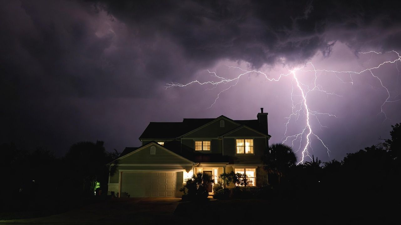 Lightning strikes behind a two-story house at night; dark storm clouds fill the sky.
