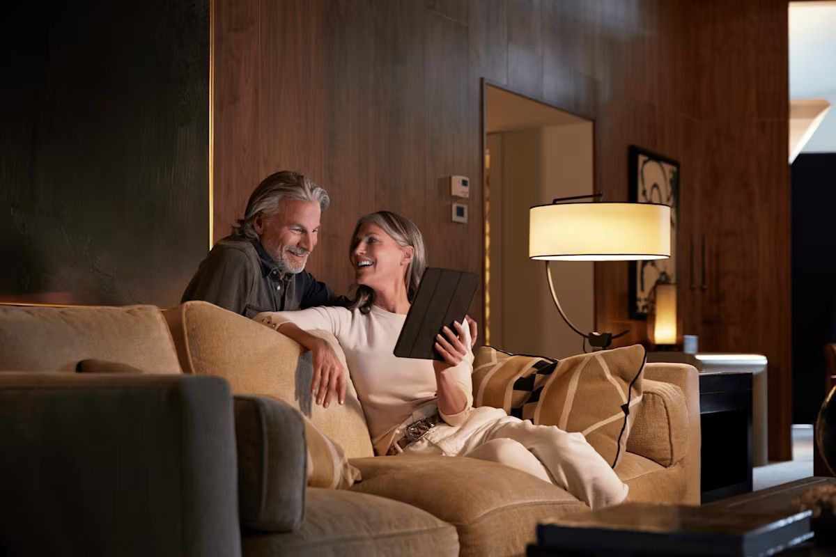 Couple smiling, looking at a tablet together on a beige sofa in a warmly lit living room.