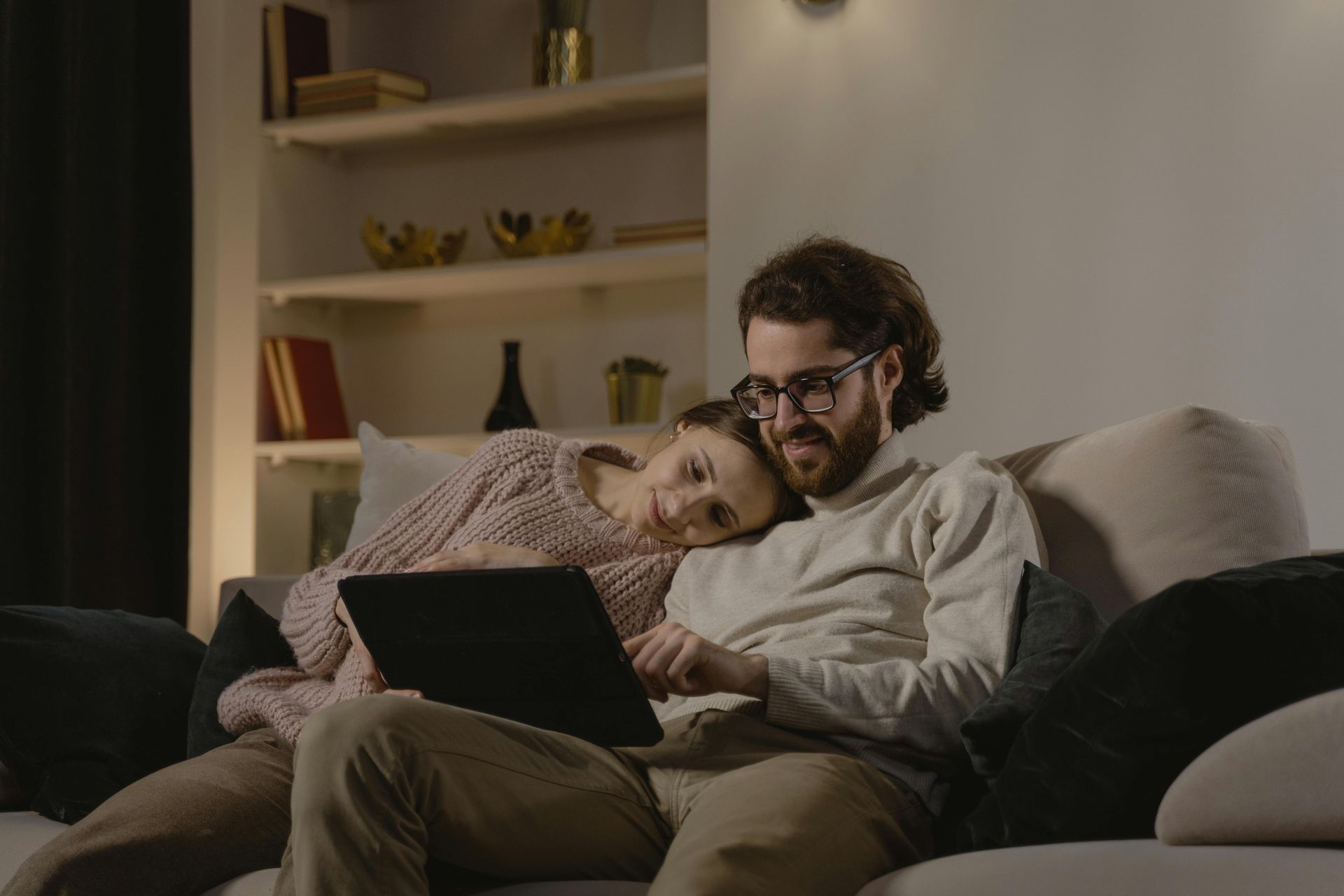 Couple on a couch looking at tablet together. Woman rests head on man's shoulder. Room with soft lighting.