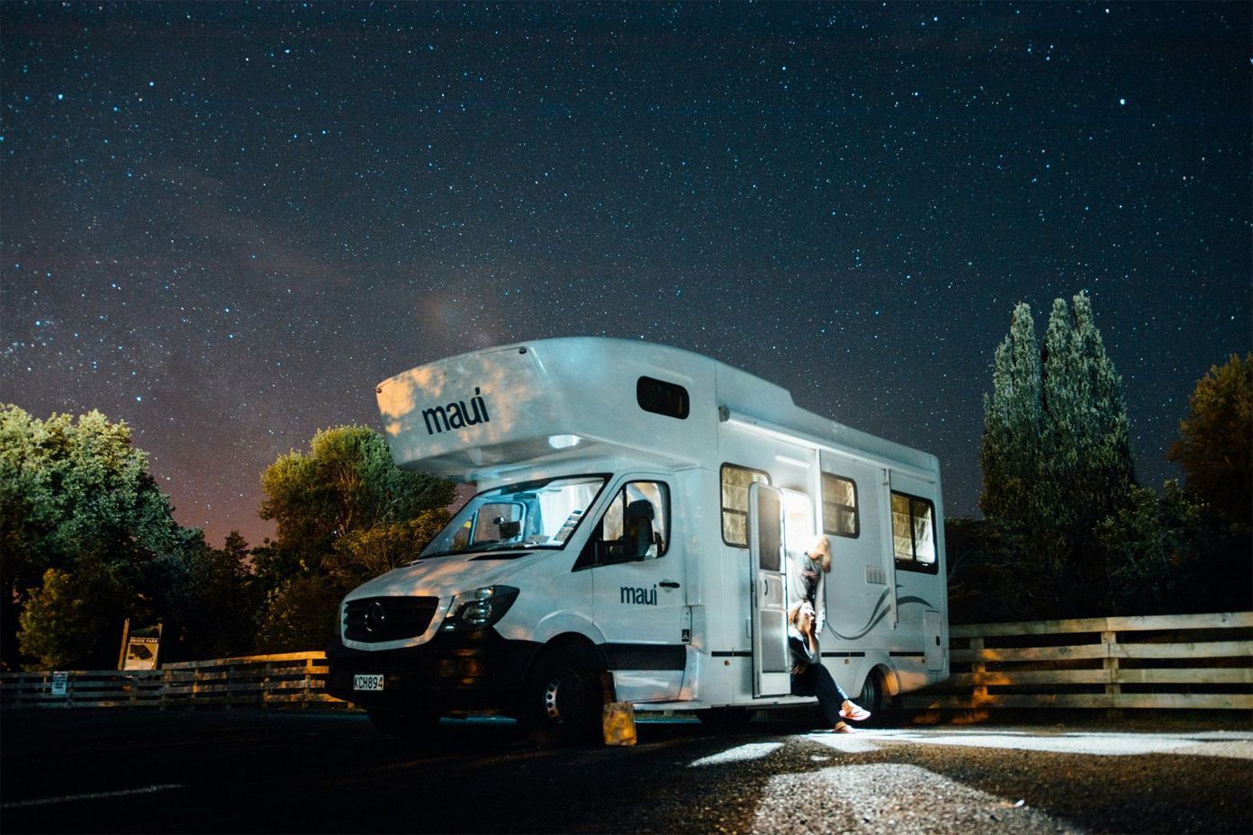 A white motorhome parked at night under a clear starry sky, with a person sitting on the open door frame.