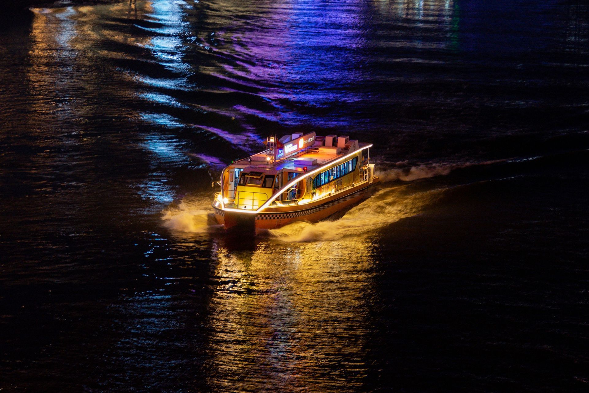 A lit tour boat travels across dark water at night, creating a golden wake against blue-toned reflections.