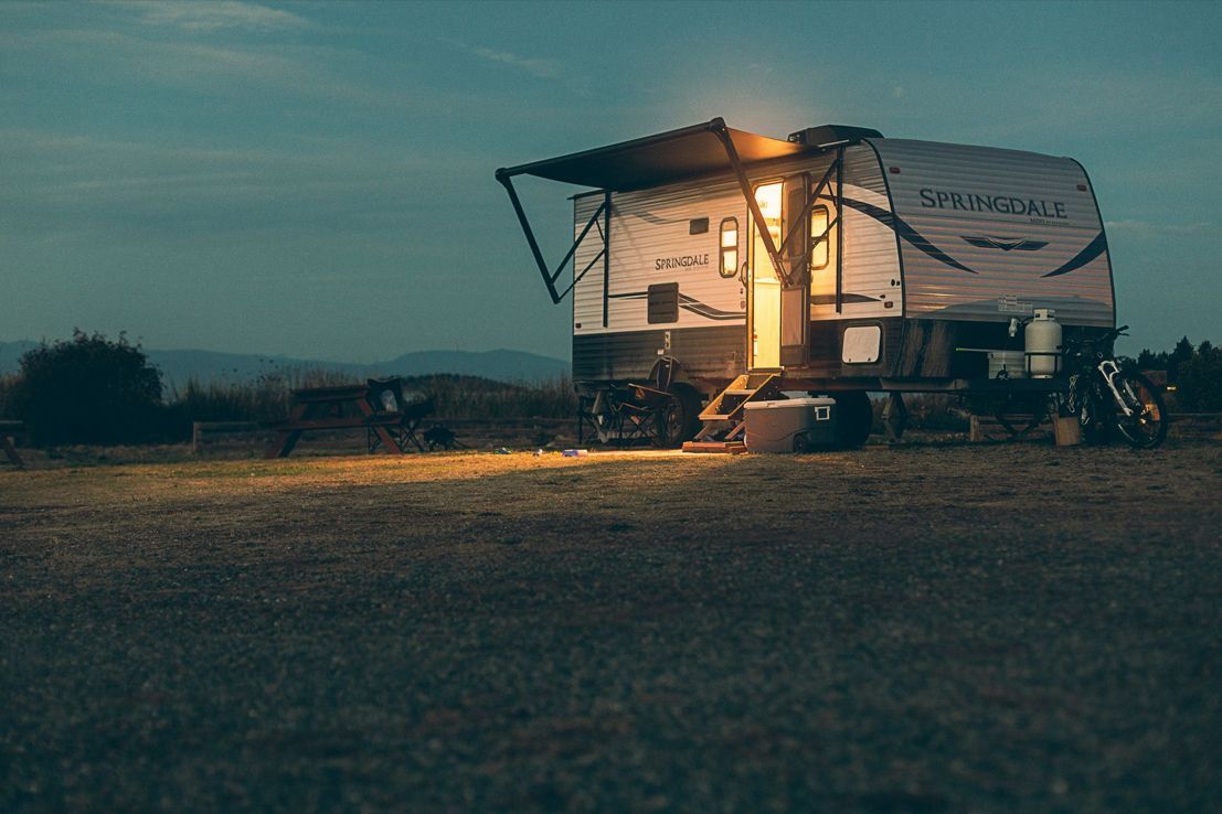 A travel trailer with its awning extended and door light on sits in a dark, open field at twilight.