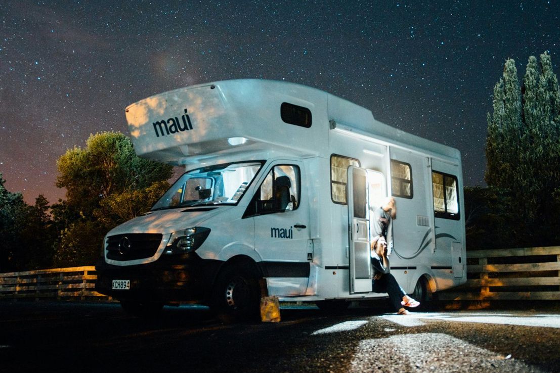 A white Maui campervan parked outdoors at night under a star-filled sky, with a person sitting by the open side door.
