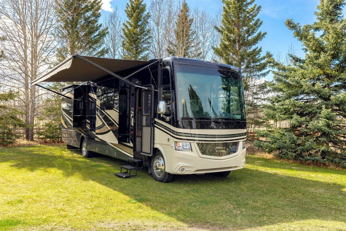 A modern, cream and black motorhome with its awning extended, parked on a grassy lawn surrounded by evergreen trees.