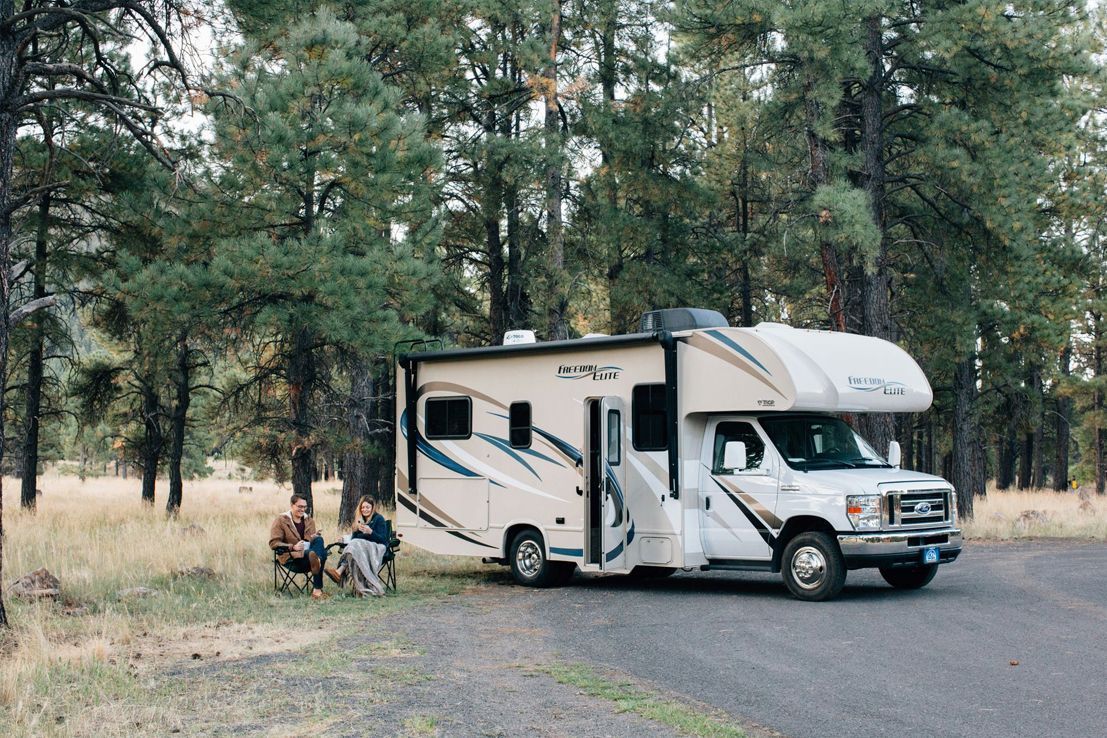 A white motorhome parked on a paved road in a pine forest, with two people sitting at a folding table nearby.