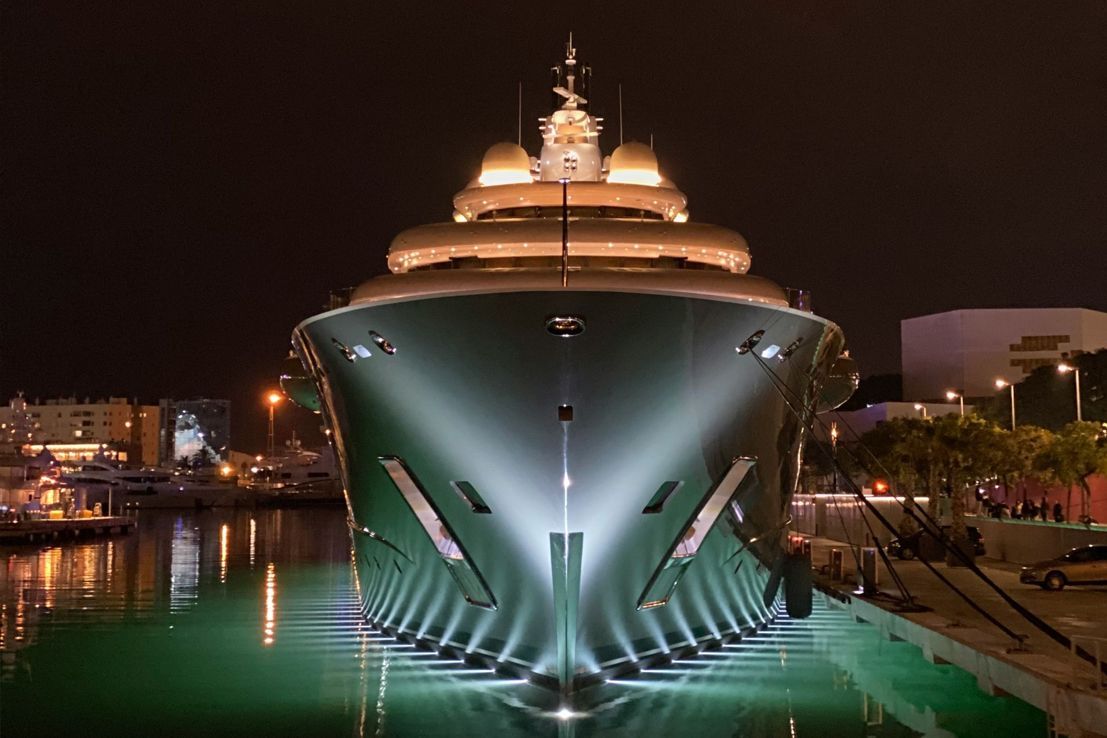 A large, dark-hulled luxury yacht docked at night, illuminated by bright floodlights reflecting on the harbor water.