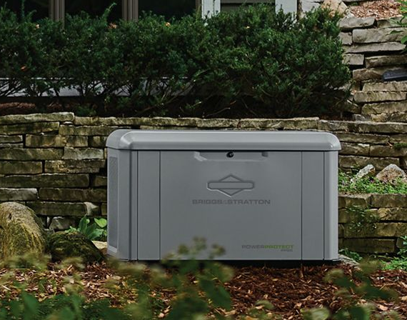 Gray Briggs & Stratton standby generator outside, against a stone wall and shrubs.
