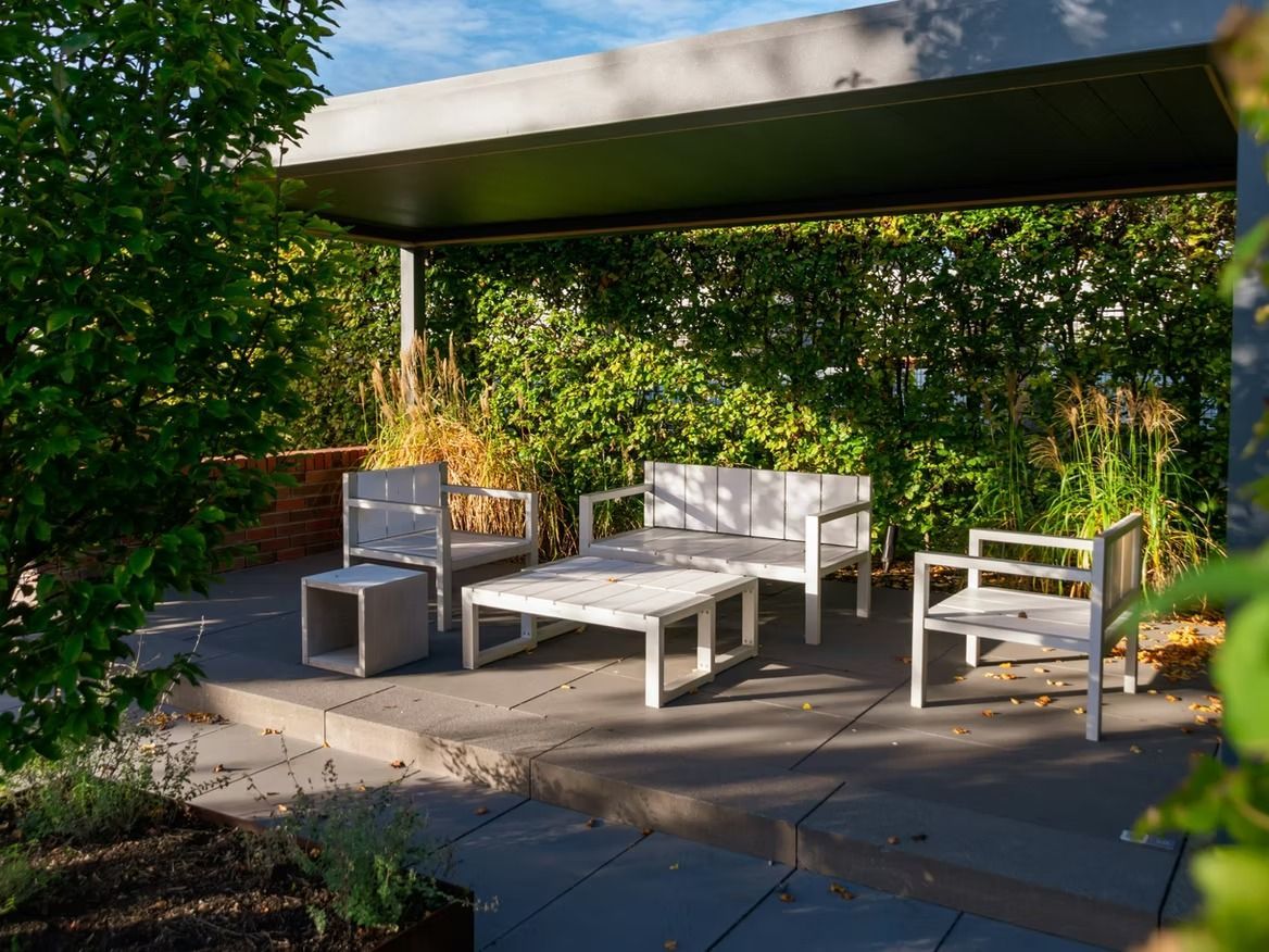Patio furniture under a modern gray canopy, surrounded by lush green foliage.
