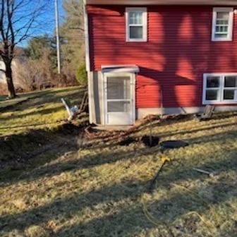 Red building with a white door and windows. Trench dug in the grassy yard.