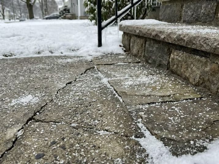 Snow-covered cracked concrete steps and walkway leading to a stone wall entrance in a snowy outdoor setting.