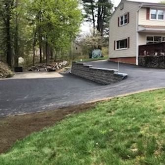 Asphalt driveway leading to a two-story house with a retaining wall. Green grass in the foreground.