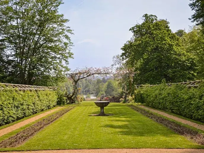 A formal garden with green lawn, hedges, a stone basin, and trees under a clear sky.