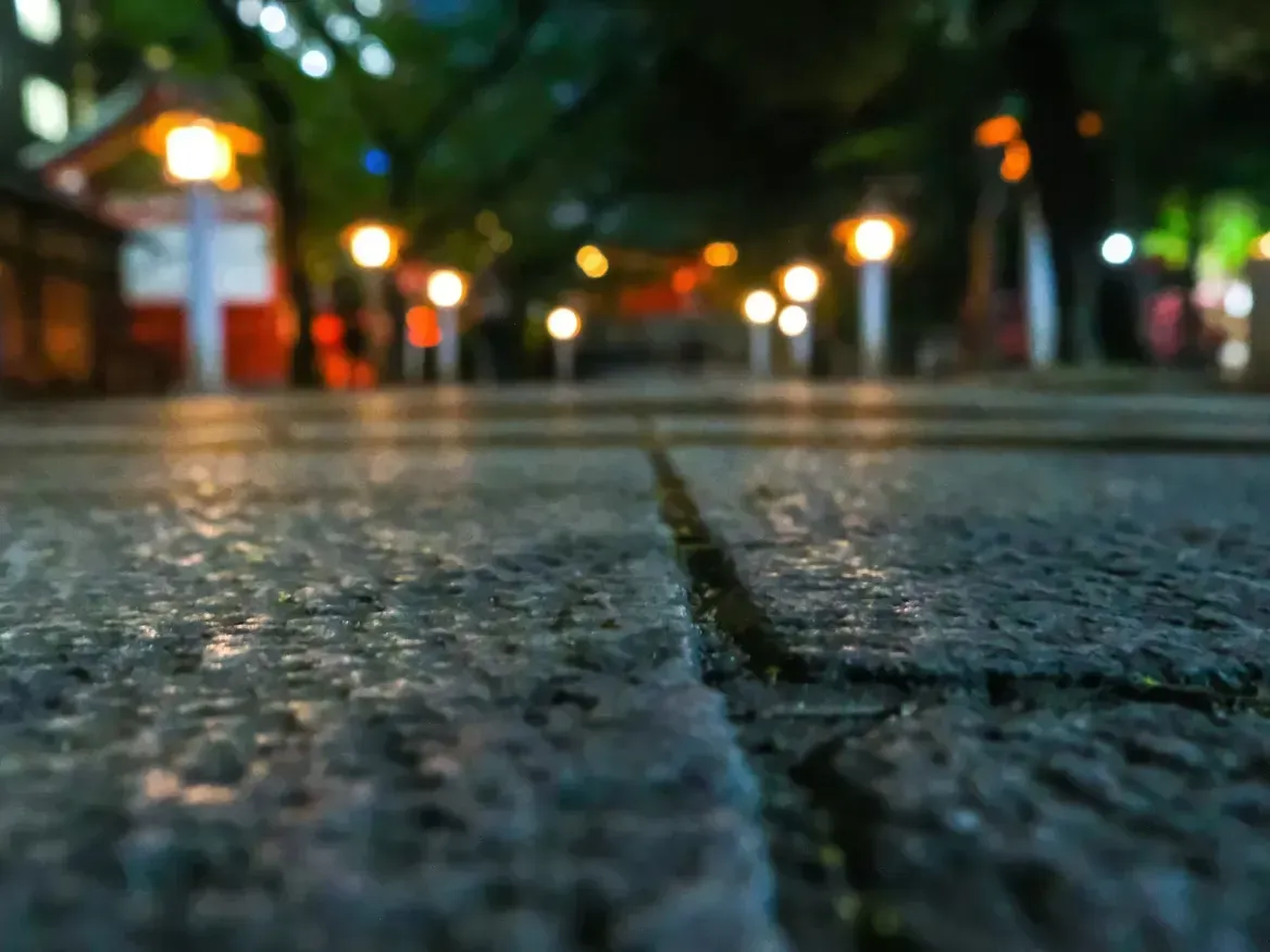 Stone path at night, lined with glowing lanterns leading to a red structure, trees on either side, blurred background.