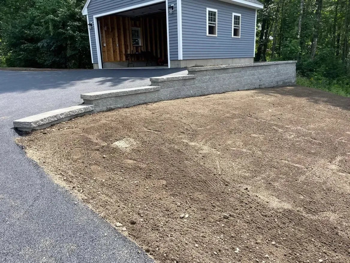 Asphalt driveway with retaining wall, dirt patch, and gray garage.