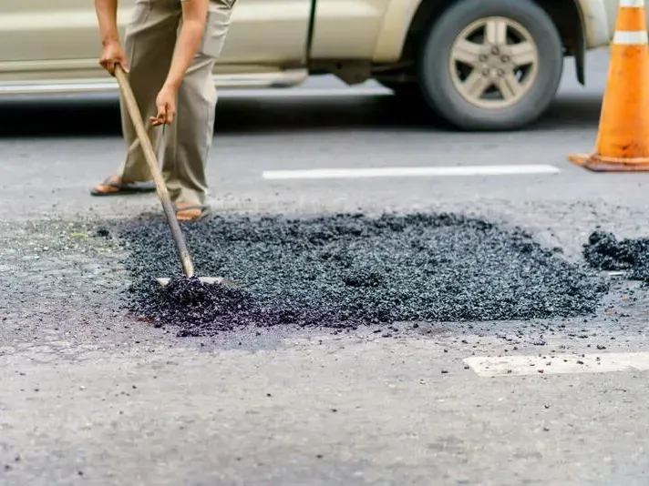 Person shovels asphalt patch on a road near a white pickup truck. An orange cone is in the background.