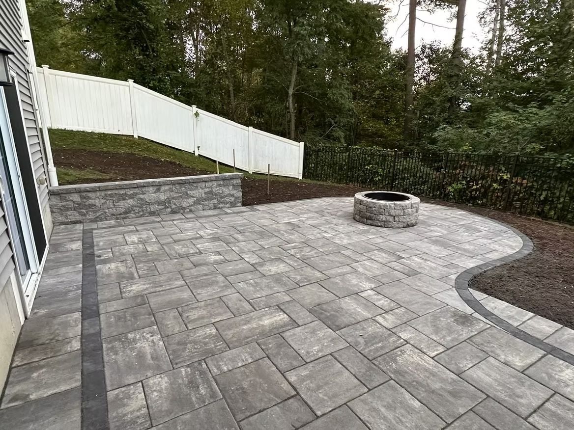 Paver patio with fire pit, stone wall, and white fence in a backyard setting.