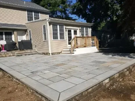 Backyard patio with gray pavers and a raised concrete border, adjacent to a beige house with a sunroom and wooden deck.