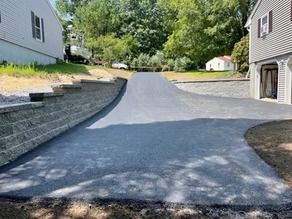 A freshly paved asphalt driveway leading between two houses, bordered by newly constructed gray stone retaining walls.