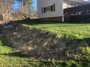 Grassy hill next to a house with exposed soil and dead grass, in a sunny outdoor setting.