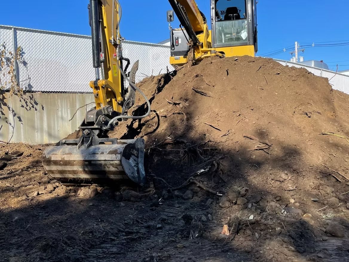 Yellow excavator digging into a pile of dirt, next to a chain-link fence.