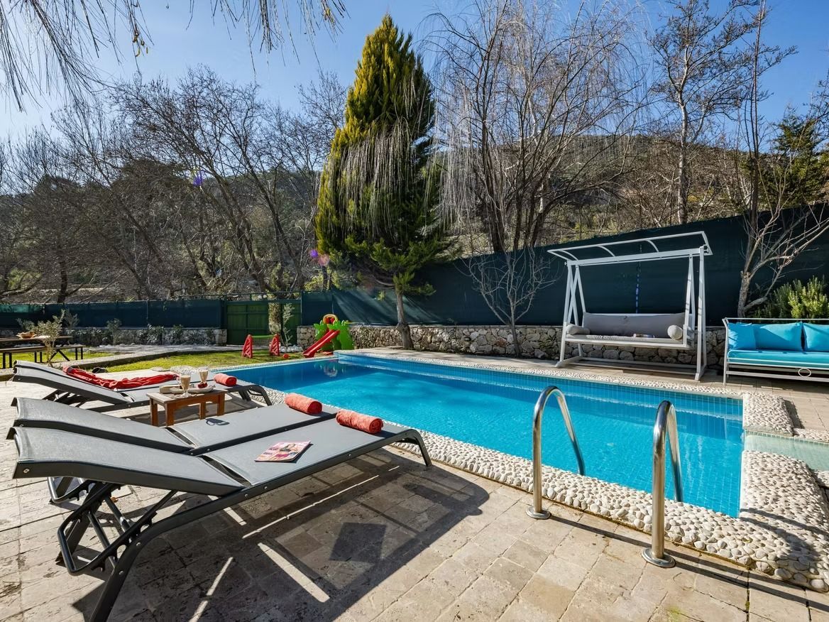 Poolside scene with a blue pool, lounge chairs, swing, and trees in the background.