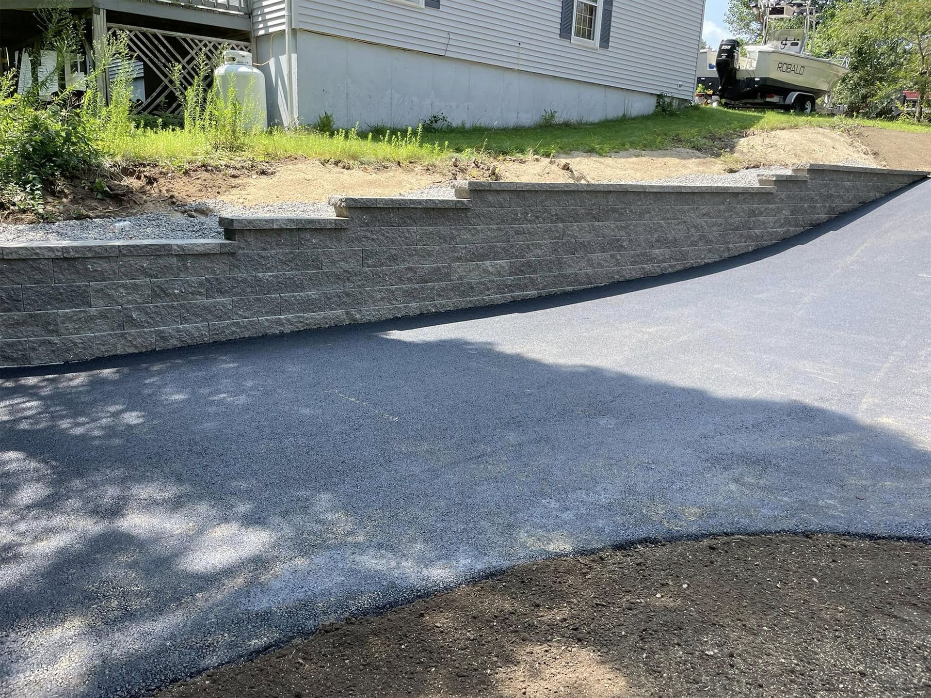 Asphalt driveway and retaining wall with steps leading uphill, grass, house in the background.