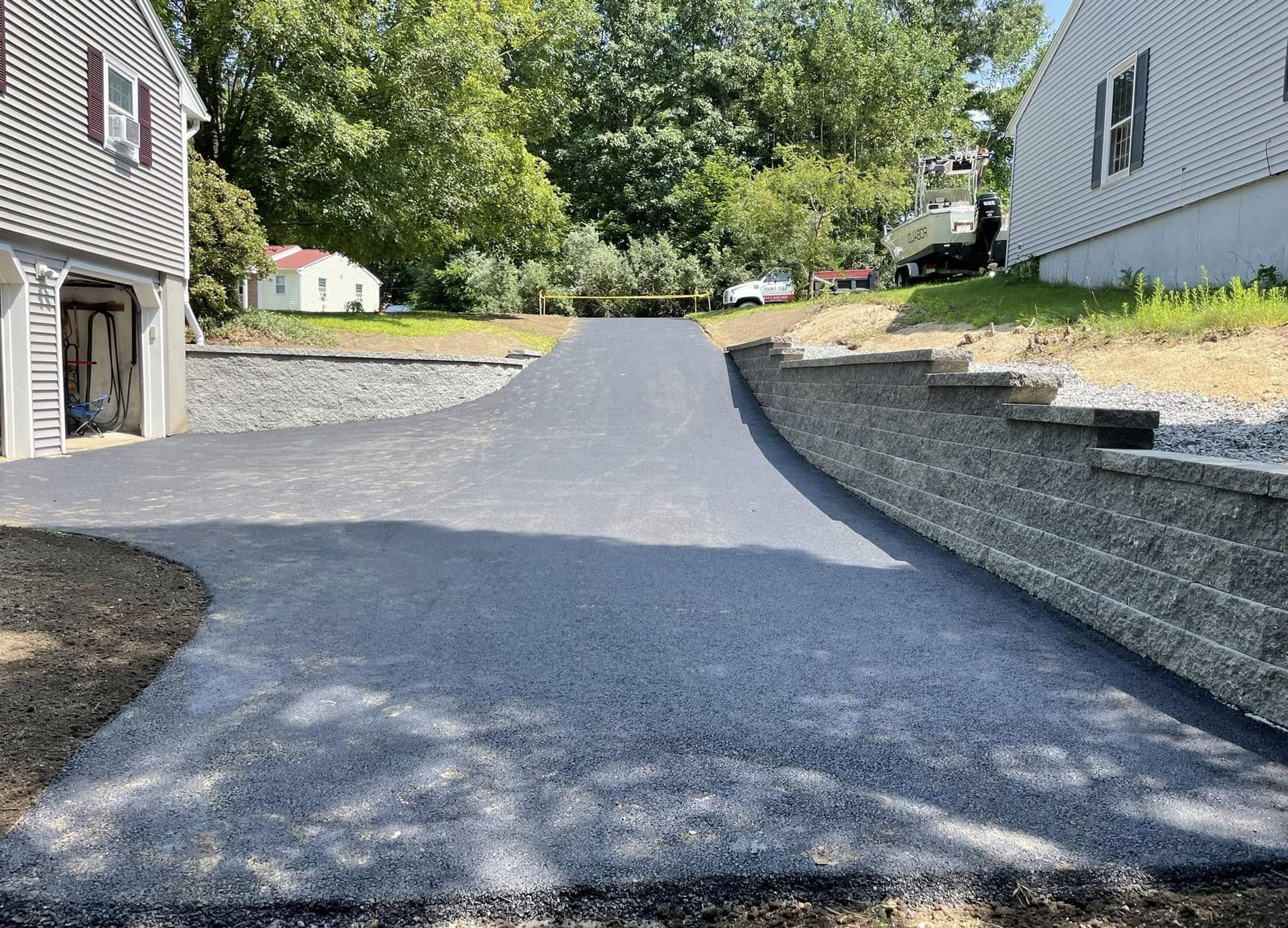Newly paved asphalt driveway curving uphill between two houses with retaining walls and grass.