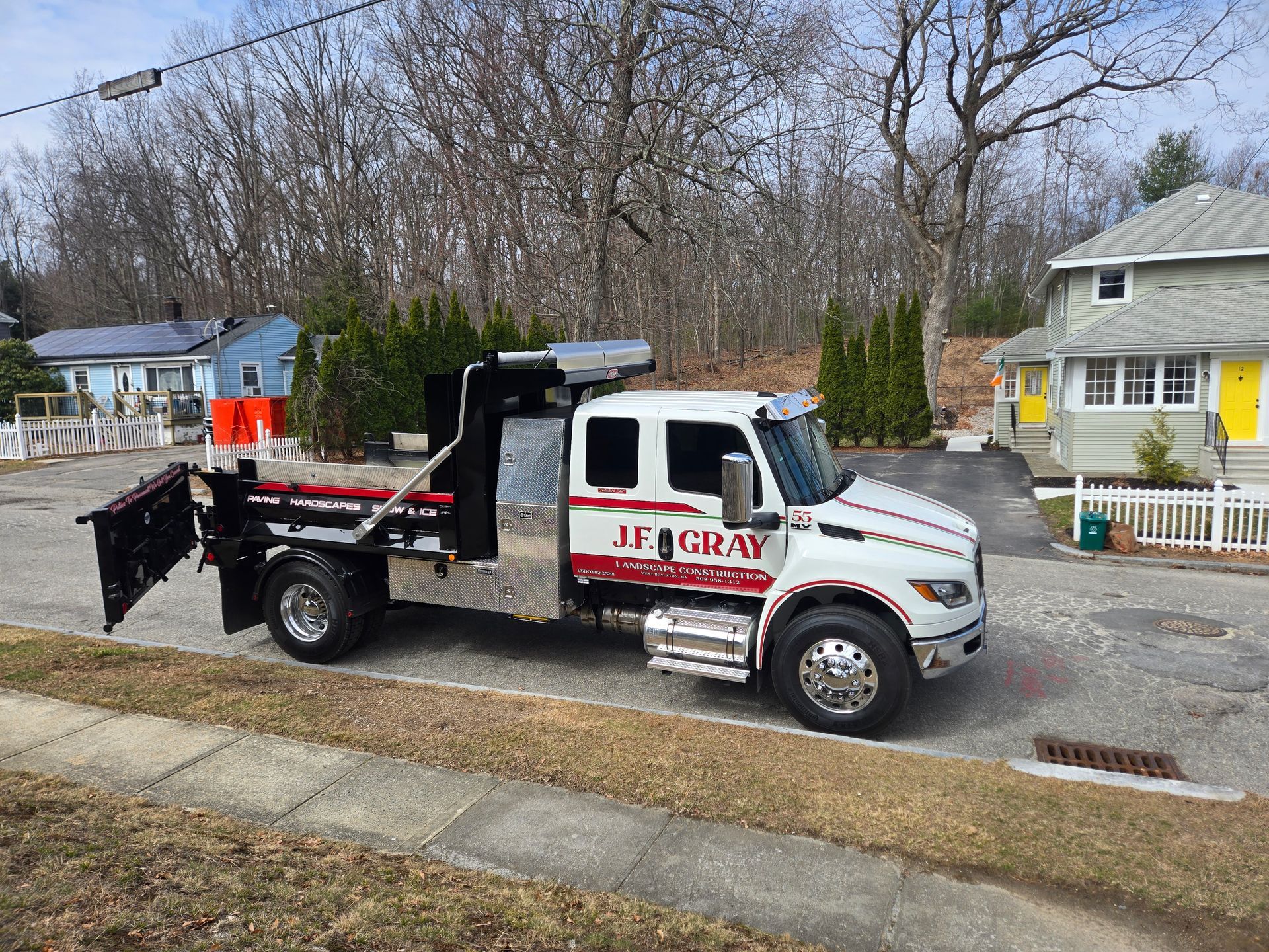 A white J.F. Gray service truck with a black flatbed and attached snow plow parked on a residential street.