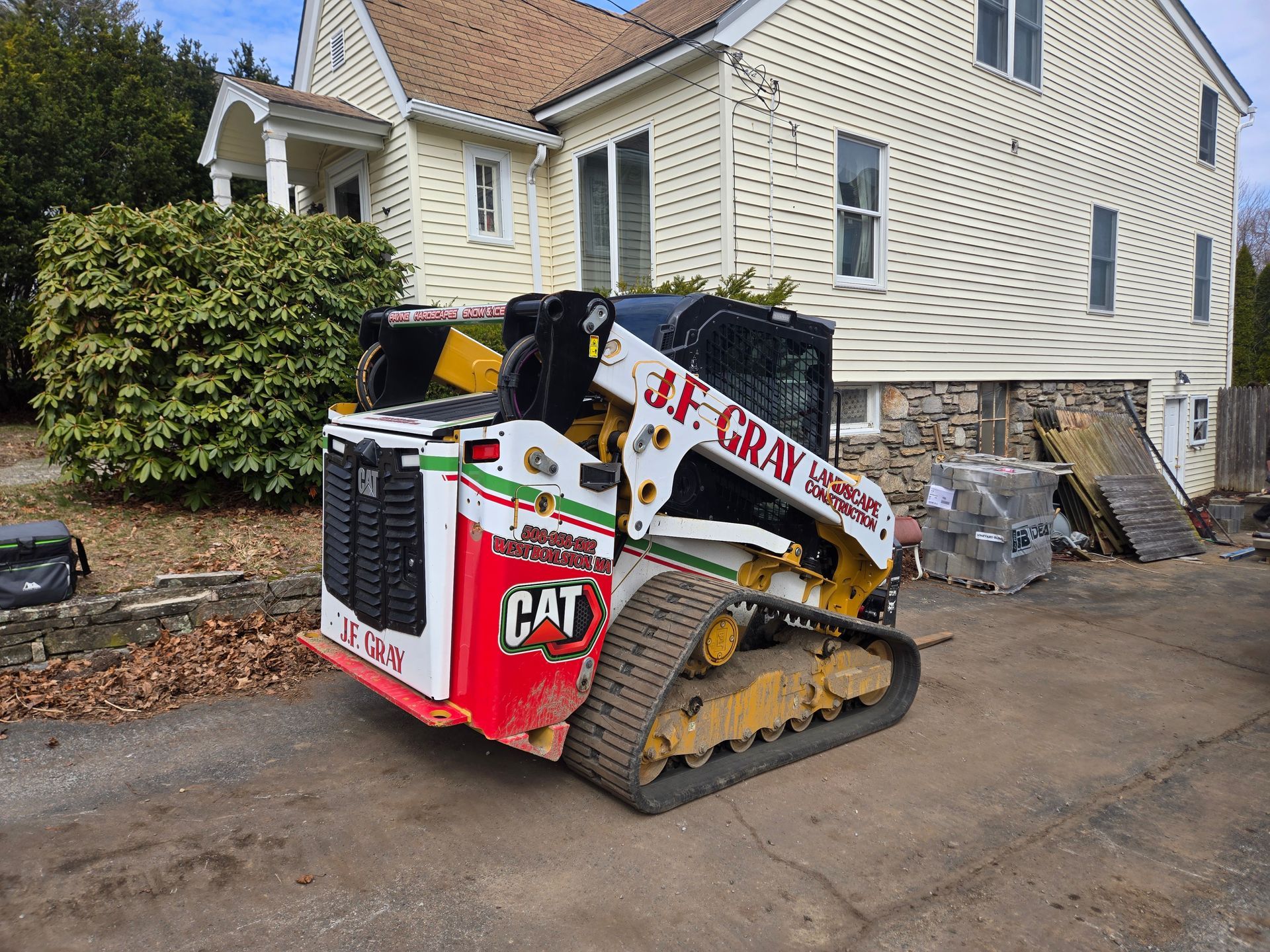 A J.F. Gray CAT compact track loader parked on a paved driveway in front of a yellow house.