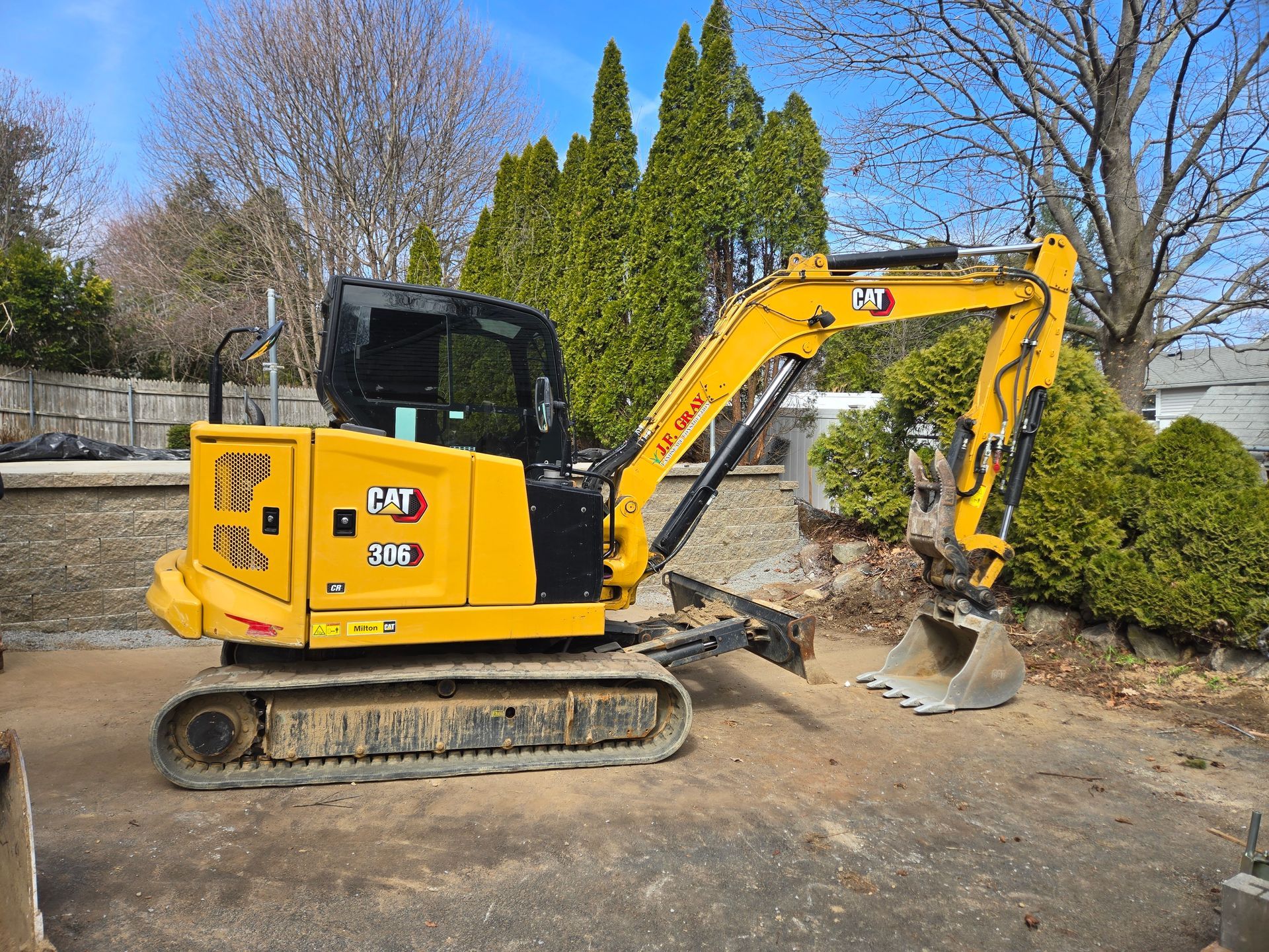 A yellow Caterpillar 305 mini excavator parked on a dirt lot outdoors with trees and a fence in the background.