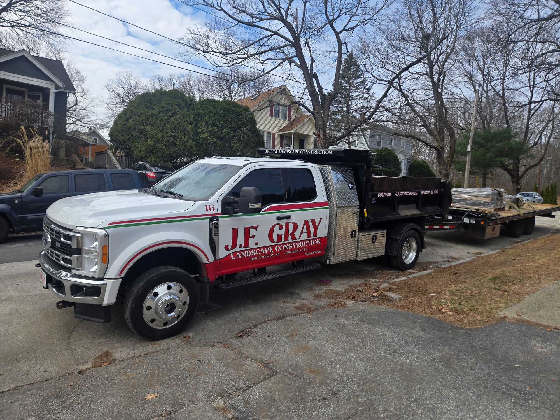 A white Ford heavy-duty pickup truck with J.F. Gray branding, towing a flatbed trailer on a suburban street.