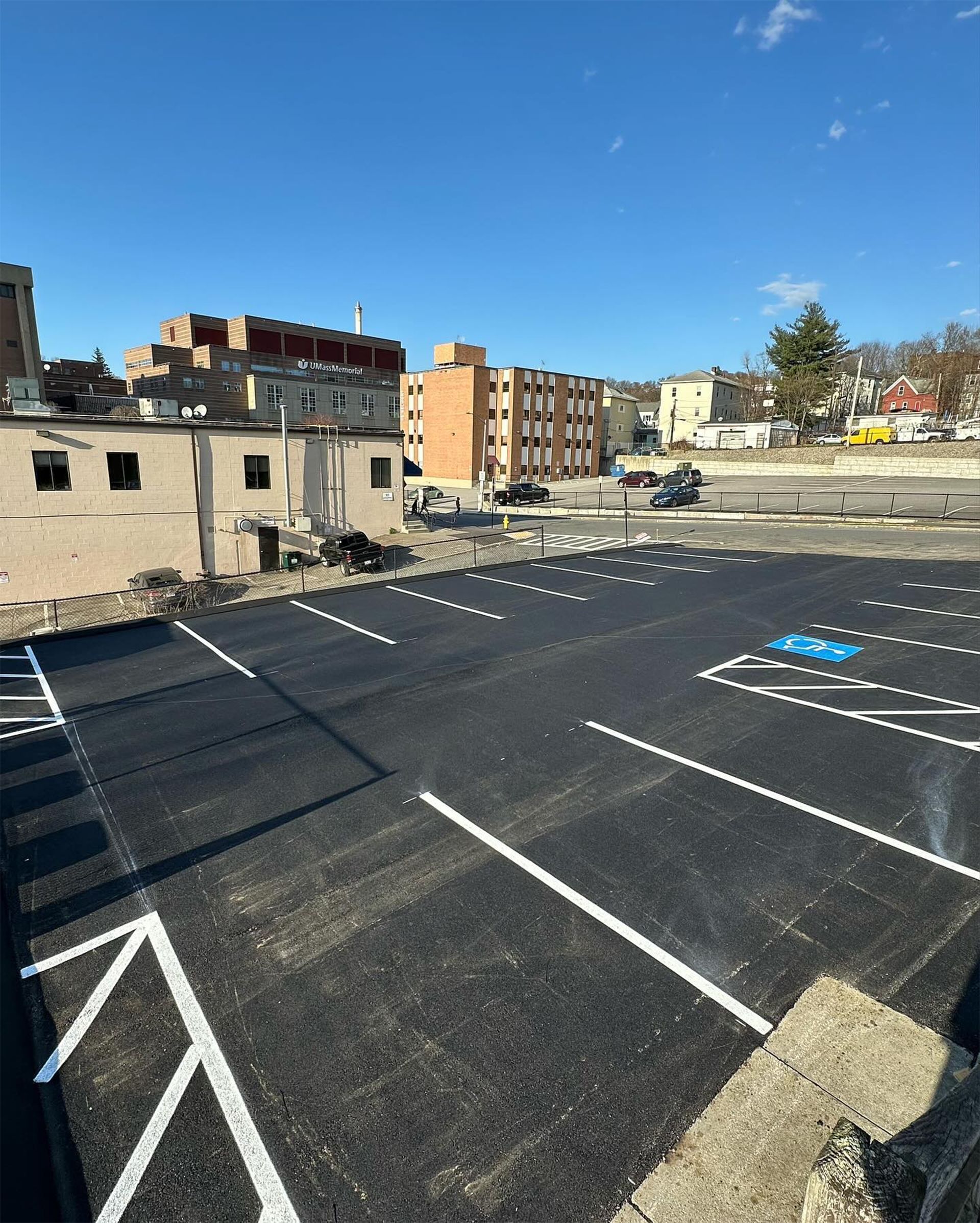 Newly paved asphalt parking lot with white painted parking space lines. Buildings in the background.