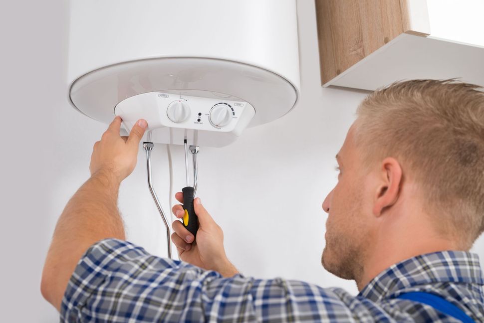 A technician in a plaid shirt uses a screwdriver to repair the control panel on the bottom of a wall-mounted water heater.