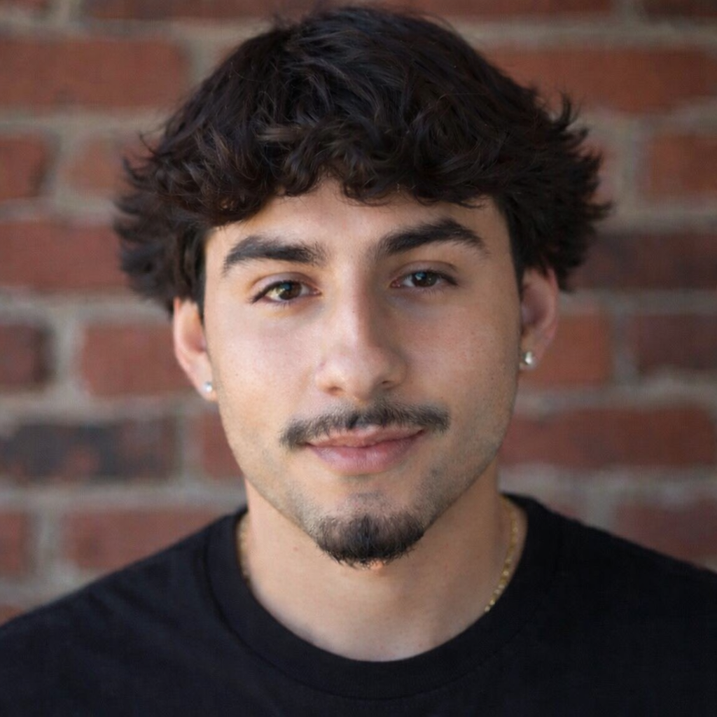 Man with dark curly hair and goatee smiling in front of a brick wall.