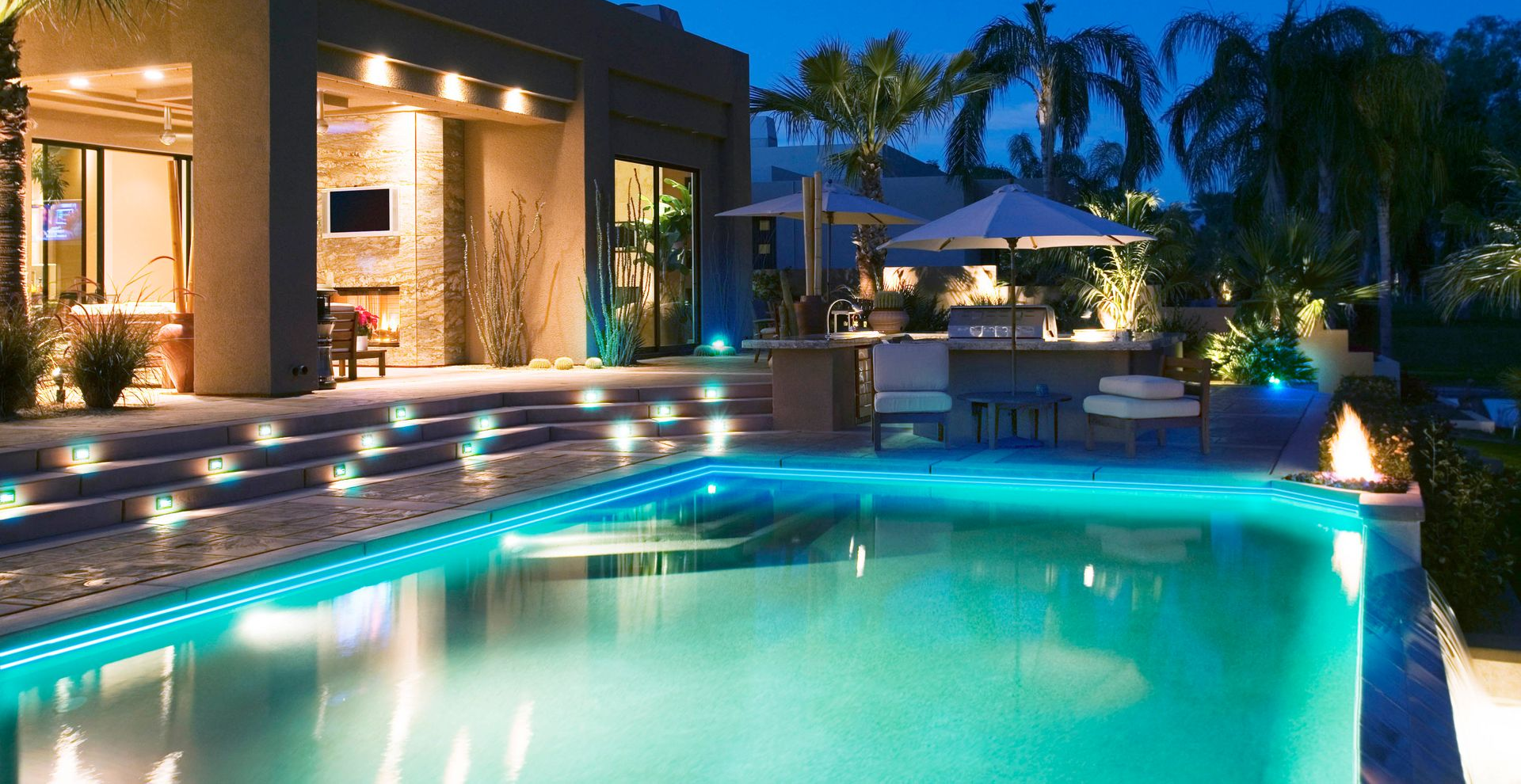 Luxury backyard pool lit at night beside a modern house and palm trees