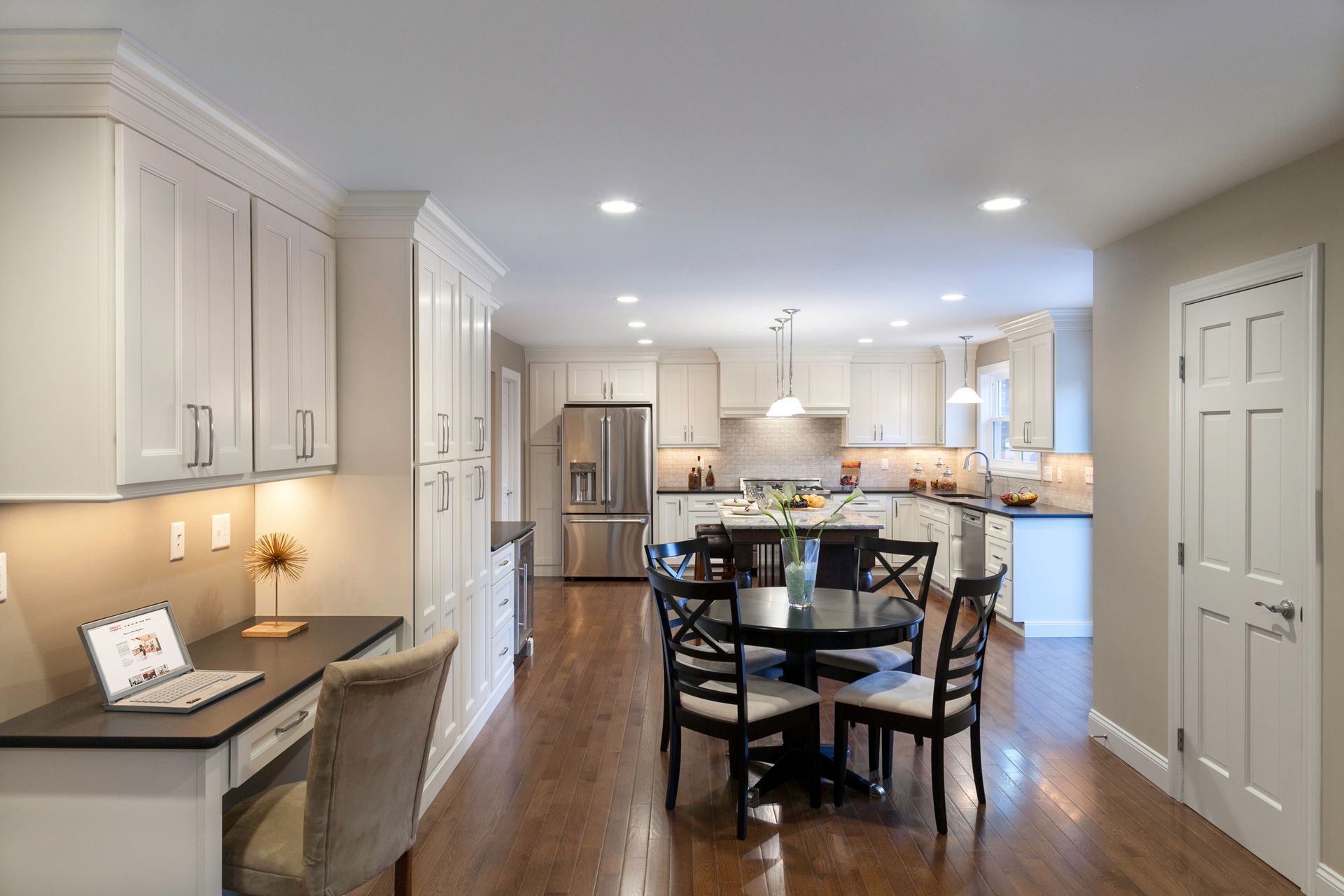 A kitchen with a table and chairs and a desk with a laptop on it.