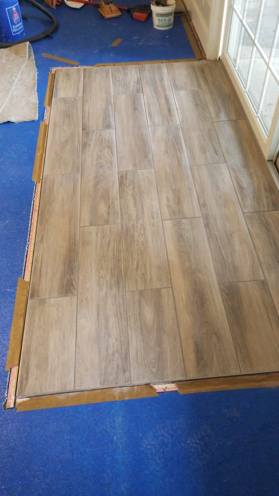 Floor tiling in progress; wood-look tiles laid on a blue floor; tile spacers in place.