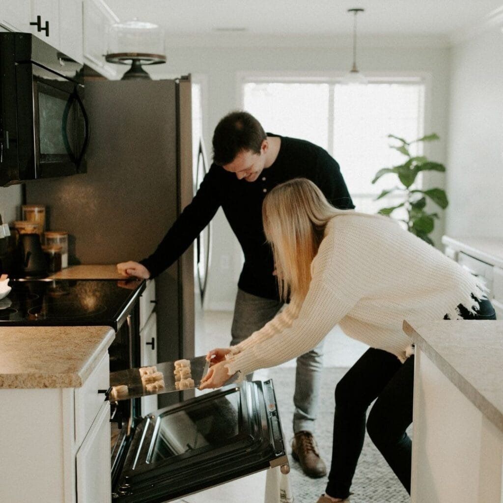Couple in kitchen looking at baked goods in the oven. The woman is putting in a tray of cookies.